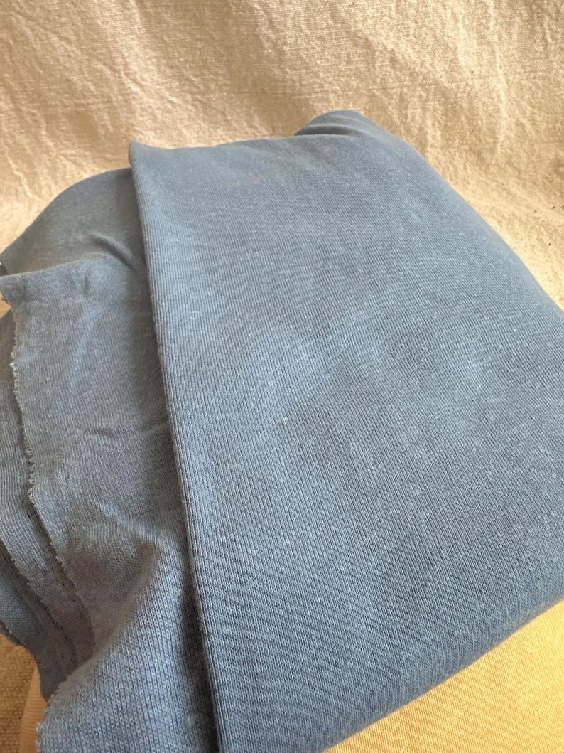 May include: A folded piece of dusty blue fabric with a visible knit texture. The material has a soft, matte appearance with subtle variations in color across the surface, showing a raw, unfinished edge on the left side of the textile.