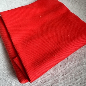 May include: A vibrant red scarf featuring a distinct ribbed knit texture. The fabric displays a consistent vertical pattern throughout the entire surface, showcasing a bright, solid color that is folded to reveal its layered construction.