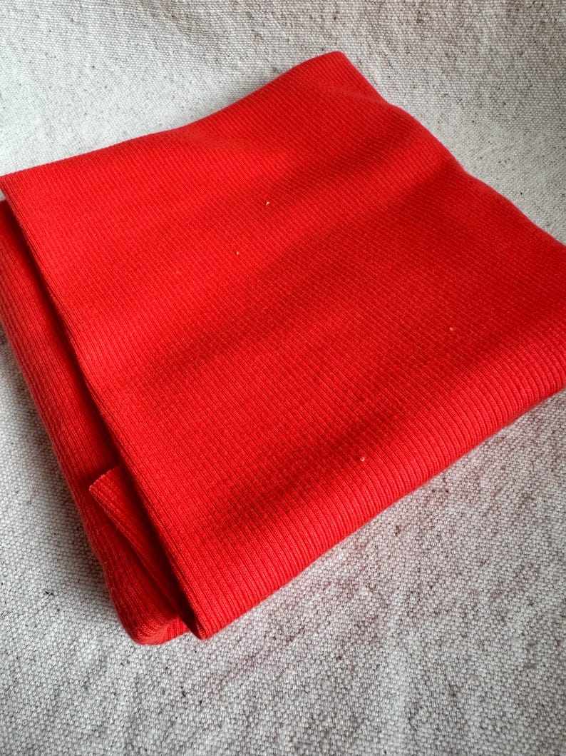 May include: A vibrant red scarf featuring a distinct ribbed knit texture. The fabric displays a consistent vertical pattern throughout the entire surface, showcasing a bright, solid color that is folded to reveal its layered construction.