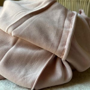 Könnte beinhalten: Nahaufnahme eines gefalteten, hellrosa Stoffes. Der Stoff hat eine subtile Textur und einen glatten, fertigen Rand. Die Falten erzeugen Schatten und Lichteffekte, die die weiche Textur des Materials hervorheben. Der Gesamteindruck ist von Komfort und Eleganz geprägt.