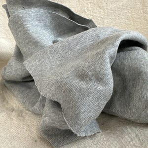 May include: A piece of heather gray knit fabric with a visible ribbed texture and raw, unfinished edges. The material is draped in soft folds, showcasing the consistent gray color and the fine, interlaced pattern of the knitted textile construction.