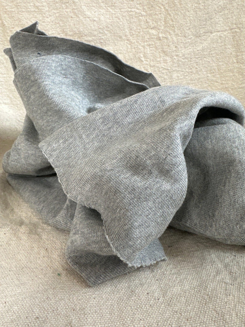 May include: A piece of heather gray knit fabric with a visible ribbed texture and raw, unfinished edges. The material is draped in soft folds, showcasing the consistent gray color and the fine, interlaced pattern of the knitted textile construction.