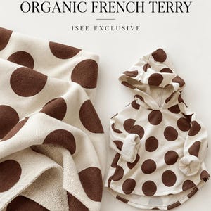 May include: A cream-colored hooded sweatshirt and a matching fabric piece, both featuring a brown polka dot pattern. The text reads: AMERICAN MILLED, OVERSIZED ORGANIC FRENCH TERRY, ISEE EXCLUSIVE. The fabric has a visible textured surface.