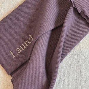 May include: A mauve-colored ribbed fabric headband featuring a twisted design. The word Laurel is printed in a light beige font on the left side of the fabric. The material has a distinct vertical ribbed texture throughout the entire surface.