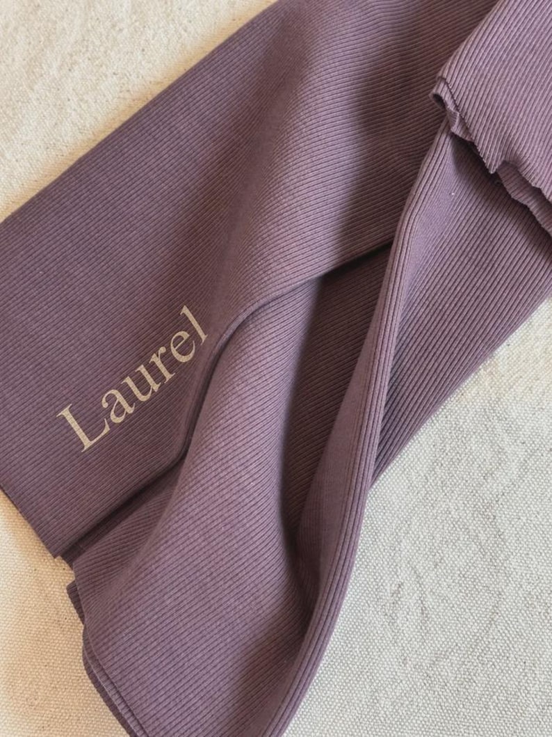 May include: A mauve-colored ribbed fabric headband featuring a twisted design. The word Laurel is printed in a light beige font on the left side of the fabric. The material has a distinct vertical ribbed texture throughout the entire surface.