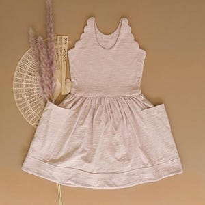 May include: A pale pink dress featuring a scalloped neckline and pockets. The dress has a gathered skirt and is made of a soft, textured fabric. A decorative fan and dried flowers are arranged to the left of the dress.
