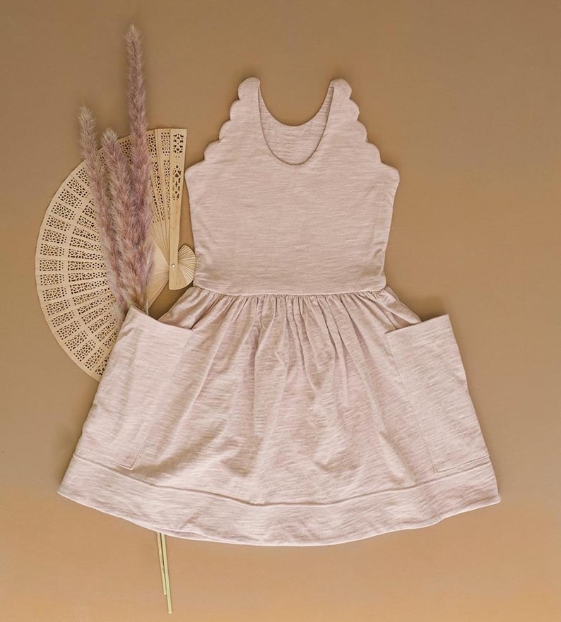 May include: A pale pink dress featuring a scalloped neckline and pockets. The dress has a gathered skirt and is made of a soft, textured fabric. A decorative fan and dried flowers are arranged to the left of the dress.