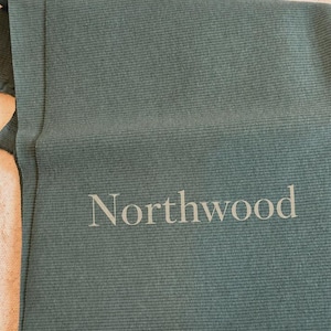 May include: A dark sage green ribbed fabric textile featuring the word Northwood printed in a light gray sans-serif font. The material has a consistent vertical ribbed texture throughout the surface, creating a linear pattern across the fabric.