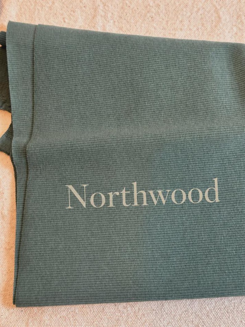 May include: A dark sage green ribbed fabric textile featuring the word Northwood printed in a light gray sans-serif font. The material has a consistent vertical ribbed texture throughout the surface, creating a linear pattern across the fabric.