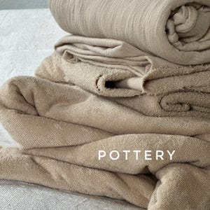 May include: A pile of beige towels, some folded and others rolled, with the word "POTTERY" in white capital letters. The towels are made of a soft, textured material.