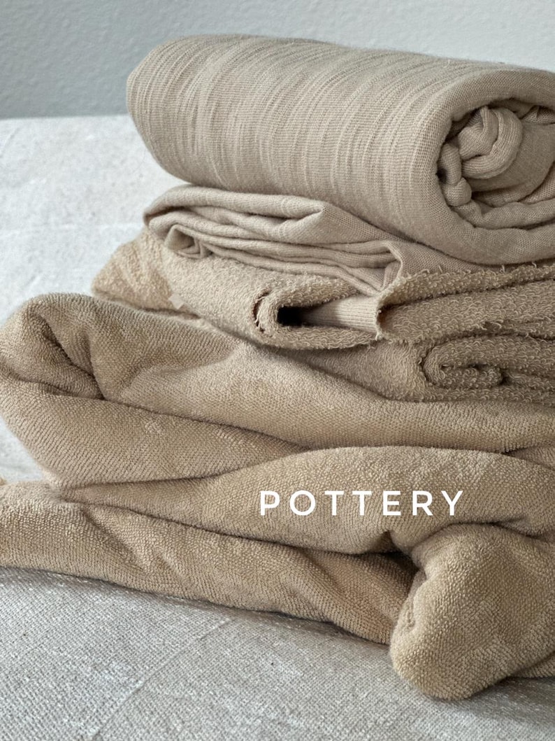 May include: A pile of beige towels, some folded and others rolled, with the word "POTTERY" in white capital letters. The towels are made of a soft, textured material.