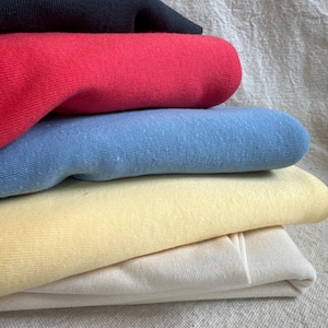 May include: A stack of folded sweaters in various colors: black, red, light blue, yellow, and cream. The sweaters are made of a soft, knitted material and are neatly arranged on top of each other. The background is a neutral-colored fabric.