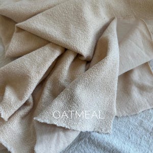 Puede incluir: Primer plano de una tela beige con superficie texturizada. La tela está doblada y drapeada, con la palabra "OATMEAL" impresa en blanco en el centro. La tela parece suave y ligera.