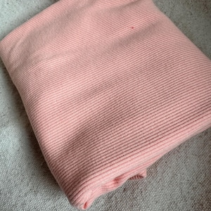 May include: A folded piece of light pink fabric featuring a consistent vertical ribbed texture. The material is soft and pliable, showing clear parallel lines running across the surface of the textile, creating a uniform and structured appearance.