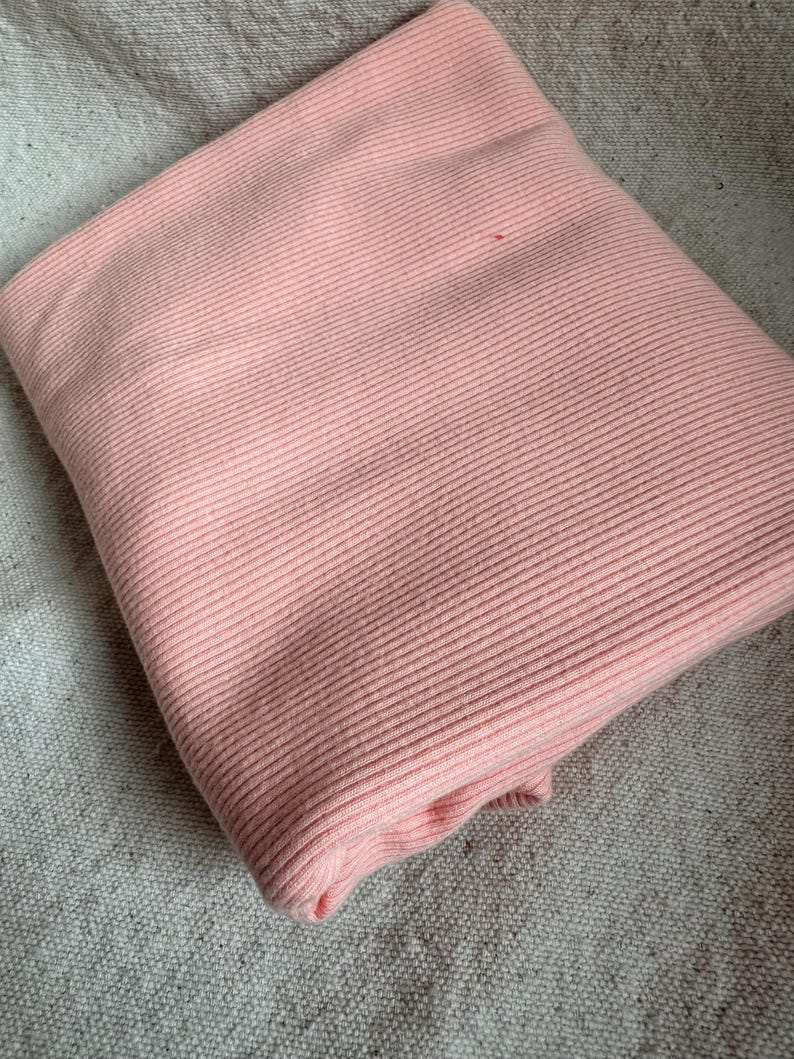 May include: A folded piece of light pink fabric featuring a consistent vertical ribbed texture. The material is soft and pliable, showing clear parallel lines running across the surface of the textile, creating a uniform and structured appearance.