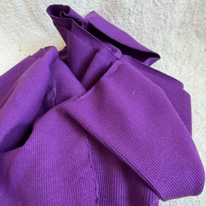 May include: A vibrant purple long-sleeved garment made of a ribbed knit fabric. The material features a distinct vertical texture throughout, with visible seams and folded sections that create a layered, draped appearance in the fabric.