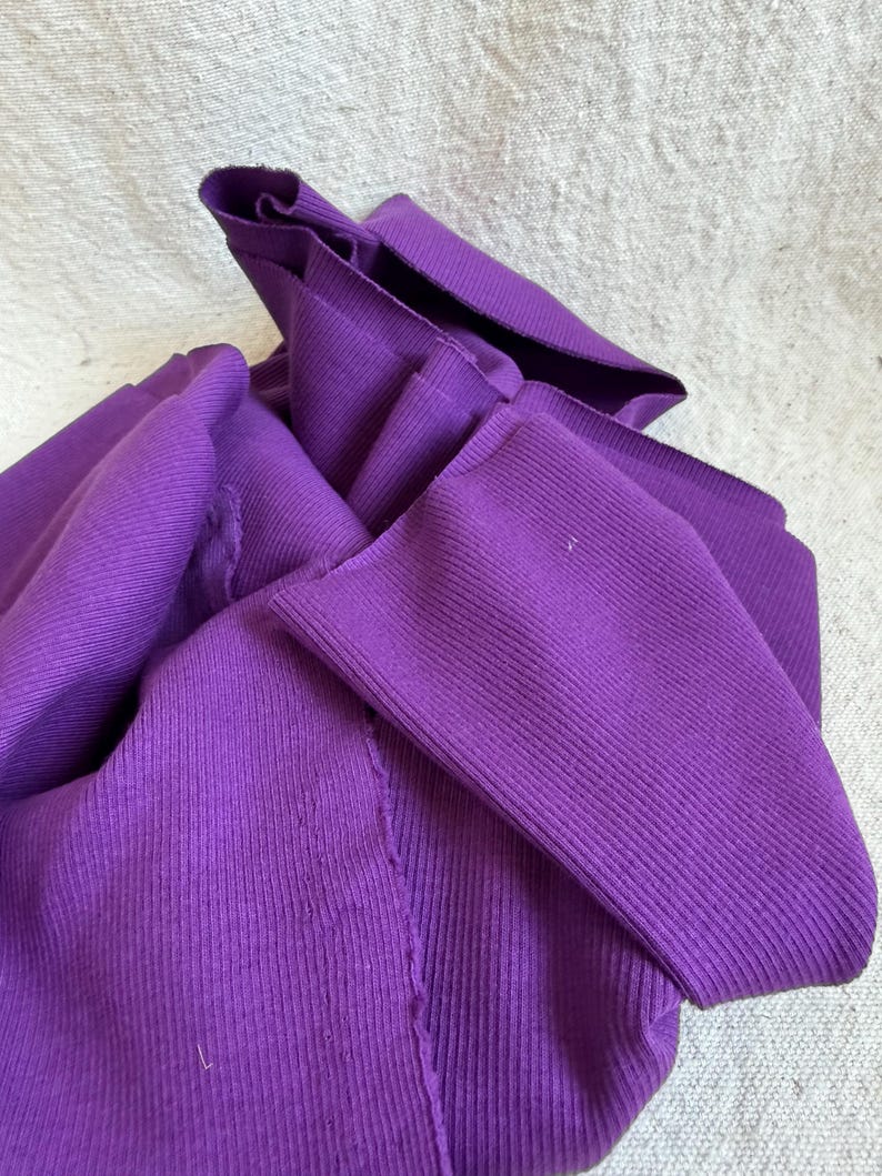 May include: A vibrant purple long-sleeved garment made of a ribbed knit fabric. The material features a distinct vertical texture throughout, with visible seams and folded sections that create a layered, draped appearance in the fabric.