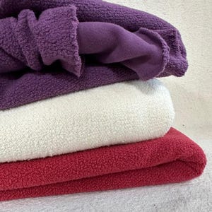 Può includere: Una pila di tessuto in pile piegato nei toni del viola, bianco e rosso. Lo strato superiore è di un viola intenso, che rivela un interno viola più chiaro. Sotto, uno strato bianco, seguito da uno strato rosso vibrante, tutti con un aspetto strutturato.