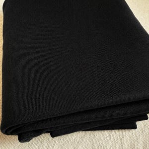 May include: A solid black textile fabric featuring a fine, consistent ribbed knit texture. The material is folded into multiple layers, showcasing the uniform vertical lines and the deep, matte black color of the woven yarn throughout the piece.