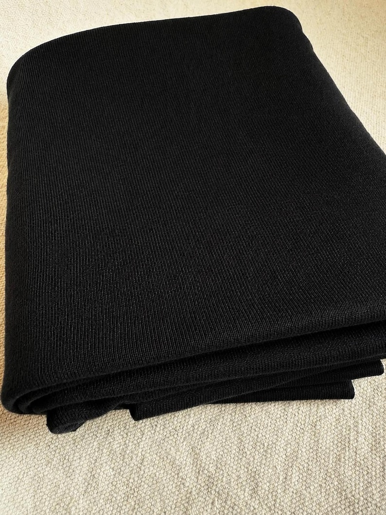 May include: A solid black textile fabric featuring a fine, consistent ribbed knit texture. The material is folded into multiple layers, showcasing the uniform vertical lines and the deep, matte black color of the woven yarn throughout the piece.