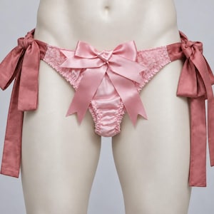Pode incluir: Uma cueca de cetim e renda rosa claro com um grande laço de cetim ao centro e fitas laterais a condizer. A peça apresenta um painel central franzido e detalhes em renda, rematada com longas e fluidas fitas de cetim em cada lado da anca.