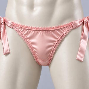 Op de afbeelding: Een lichtroze bikini-onderbroek van satijn met zijdelingse strikjes en een verfijnde, geschulpte kanten rand langs de voorzijde. De stof heeft een gladde, reflecterende afwerking en een zachte, pastelroze kleur.