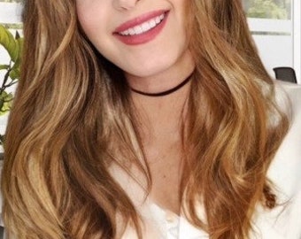 May include: A close-up of a person with long, wavy, light brown hair and a black choker necklace. The person is smiling, revealing white teeth and red lipstick. The background is blurred, with a glimpse of greenery and a window.