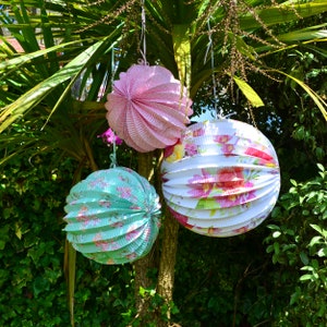 May include: Three paper lanterns hanging from a tree branch. The lanterns are round and have a floral pattern. One lantern is pink, one is white with pink flowers, and one is green with white flowers.