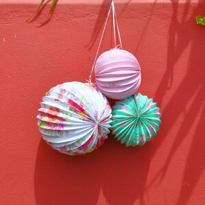 May include: Three paper lanterns hanging from a red wall. The lanterns are pink, white with floral patterns, and green with a starburst pattern.