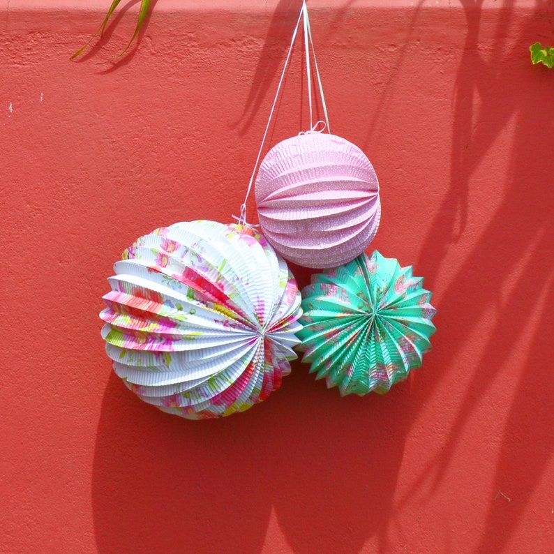 May include: Three paper lanterns hanging from a red wall. The lanterns are pink, white with floral patterns, and green with a starburst pattern.