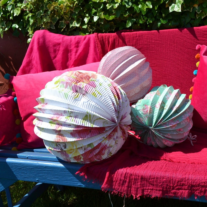 May include: Three decorative paper lanterns with floral patterns. The lanterns are white with pink, green, and blue accents. The lanterns are sitting on a red couch.