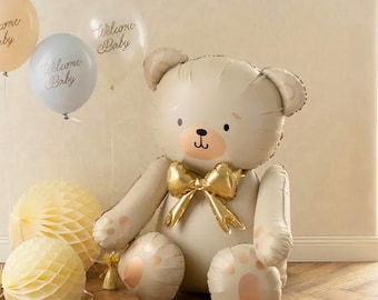 May include: A large, beige teddy bear balloon with a gold bow tie, surrounded by balloons that say "Welcome Baby" and honeycomb paper decorations. The bear has pink accents on its paws and snout.