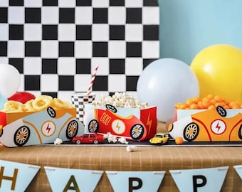 May include: Three race car themed party favor boxes filled with snacks. The boxes are red, blue, and yellow with a black lightning bolt design. The boxes are sitting on a brown table with a blue and white checkered background.