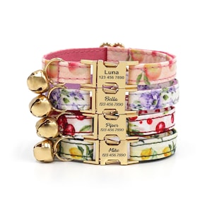 May include: A stack of cat collars with gold-coloured bells and buckles. Each collar displays a different floral or fruit pattern in pink, purple, red, and yellow. The buckles are engraved with names and phone numbers: Luna, Bella, Piper, and Mike.