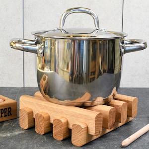May include: A stainless steel pot with a lid sits on a wooden pot holder. The pot holder is made of light brown wood and has a rectangular shape. A wooden spoon is lying on the counter next to the pot holder.