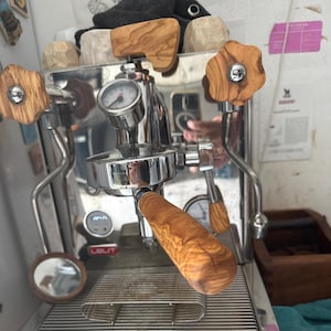 Może przedstawiać: Srebrna ekspres do kawy z drewnianymi uchwytami. Ekspres ma manometr i dyszę do spieniania mleka. Ekspres stoi na drewnianej powierzchni.