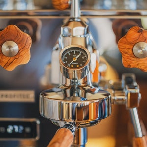 Puede incluir: Primer plano de una máquina de espresso plateada con un manómetro y pomos de madera. El manómetro indica "POEMA" y una aguja negra apunta a 10 bares.