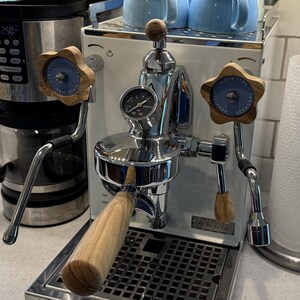 Puede incluir: Una máquina de espresso de acero inoxidable con detalles de madera y un manómetro. También se ven una cafetera de goteo y dos tazas de café azul claro. Un rollo de toallas de papel está cerca.