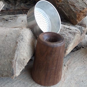 May include: A small, silver metal strainer with a perforated interior sits next to a dark brown, wooden, conical object. The objects are resting on a surface of weathered, gray wood pieces. The objects appear to be components of a smoking pipe.