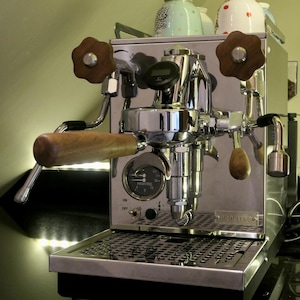 Puede incluir: Máquina de espresso de acero inoxidable con detalles en madera. Cuenta con un manómetro, un portafiltro y una lanza de vapor. Varias tazas de café están apiladas encima. La máquina mide aproximadamente 30 cm de ancho.