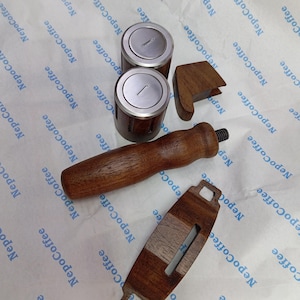 May include: A set of coffee equipment components including two cylindrical metal and wood containers, a wooden handle with a threaded metal end, a small wooden block, and a flat wooden piece with a central slot, all arranged on branded paper.