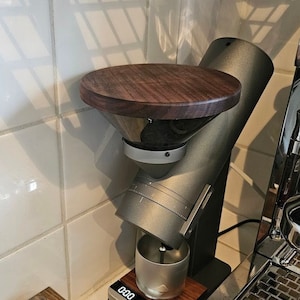 May include: A silver coffee grinder with a wooden top, a stainless steel cup, and a digital scale displaying "000 18.25". The grinder is sitting on a wooden stand with a cup holder and a tamper.