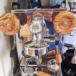 Puede incluir: Una máquina de espresso plateada con una pantalla digital que muestra "GEISINGER 8". La máquina tiene dos pomos de madera y un portafiltro con un mango de madera.