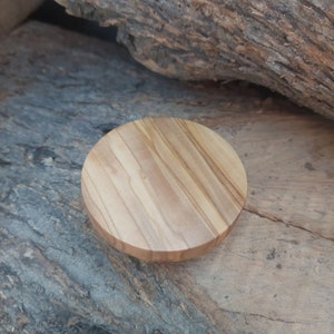 Può includere: Un coperchio rotondo in legno d'ulivo, di colore marrone chiaro con venature evidenti. Il coperchio è probabilmente per un contenitore. Lo sfondo mostra una superficie di legno marrone scuro strutturata.