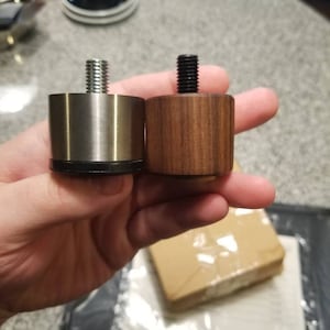Puede incluir: Dos tapas para patas de muebles, una es de metal plateado con una parte superior roscada y la otra es de madera marrón con una parte superior roscada negra.