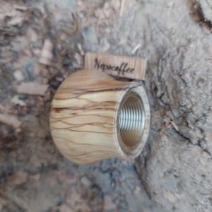 May include: A wooden coffee tamper with a brass threaded insert. The tamper is made of light wood with dark brown streaks. The tamper is labeled "Nespresso" on a small wooden tag.