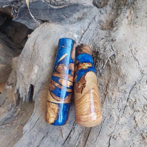 Puede incluir: Dos tapones de botella hechos a mano. Uno es una combinación de resina azul y madera, el otro es de madera natural. El tapón azul tiene un acabado brillante, mientras que el otro tiene un acabado mate. Ambos son de forma cilíndrica y se asientan sobre una superficie de madera.