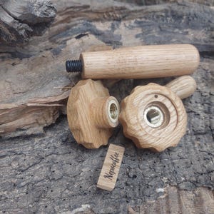 Op de afbeelding: Drie houten knoppen met schroefdraadmetalen inzetstukken. De knoppen zijn lichtbruin en hebben een getextureerd oppervlak. De knoppen zijn gerangschikt op een stuk hout met een bruine basttextuur. Een klein houten label met de tekst "Nepocoffee" is zichtbaar in het onderste midden van de afbeelding.