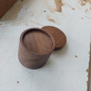 May include: A handcrafted wooden box with a lid, showcasing a natural wood grain. The box is cylindrical with a slightly tapered shape, and the lid is a flat, circular piece. The wood has a warm, brown tone, suggesting a natural finish.