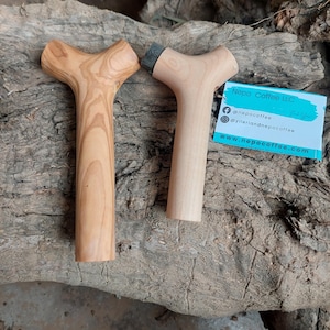 May include: Two wooden coffee tampers with a Y-shaped handle. One is light brown with a natural wood grain, and the other is a lighter color with a dark gray tamping surface. A business card with social media handles and a website is visible.